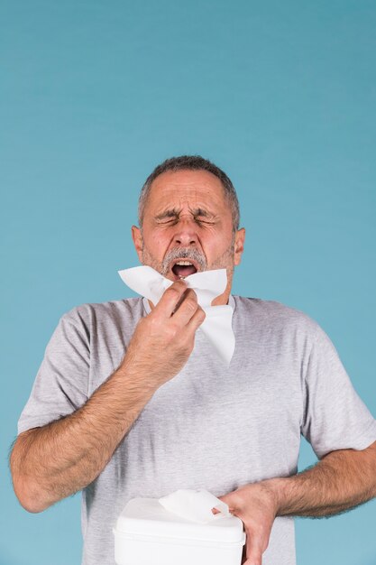 Senior man holding tissue paper about to sneeze on blue background