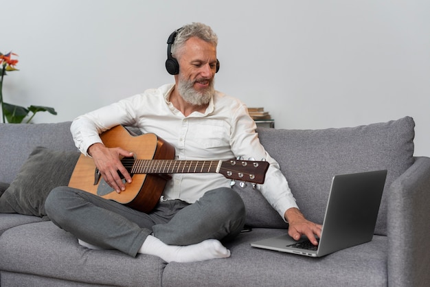 Старший мужчина дома на диване, используя ноутбук для изучения уроков игры на гитаре