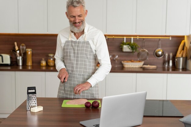 Senior man at home in the kitchen taking cooking lessons on laptop