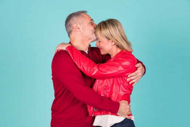 Senior man kissing on his wife's head embracing against colored background