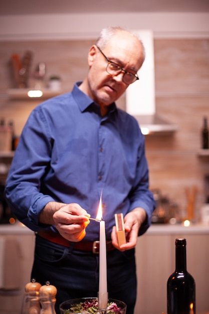 Старший мужчина зажигает свечу со спичками на кухне для романтического ужина с женой. пожилой старый муж готовит праздничную еду с здоровой пищей для празднования годовщины, сидя за столом.