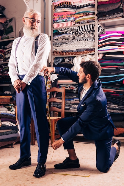 Senior man looking at male fashion designer taking measurement of his pant in the shop