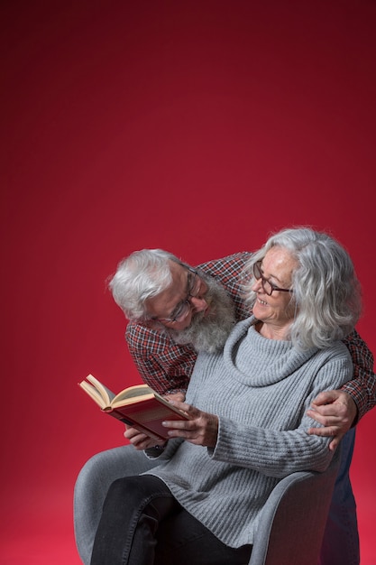 Free photo senior man loving her wife sitting on chair reading the book against red backdrop