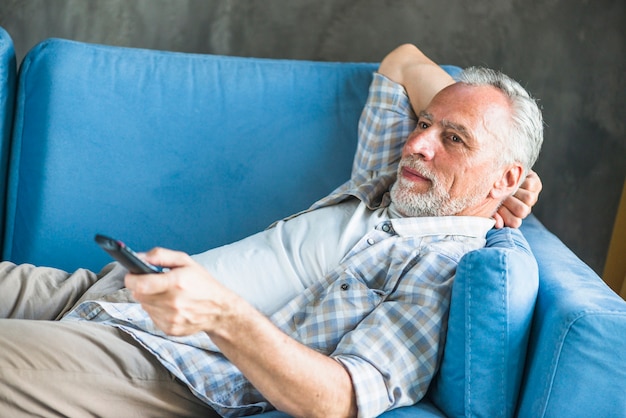 Senior man lying on blue sofa using remote control