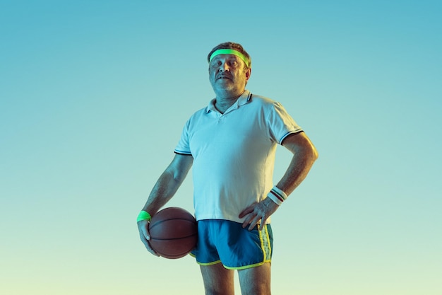 Старший мужчина играет в баскетбол на градиентном фоне в неоновом свете. Кавказский мужчина-модель в отличной форме остается активным, спортивным.