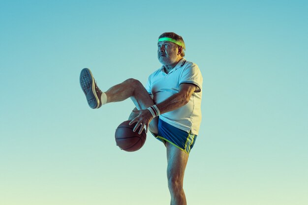 Senior man playing basketball on gradient wall in neon light. Caucasian male model in great shape stays active, sportive. Concept of sport, activity, movement, wellbeing, healthy lifestyle.