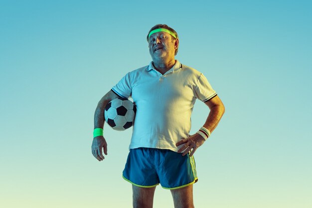 Старший мужчина играет в футбол в спортивной одежде на градиентном фоне и неоновом свете