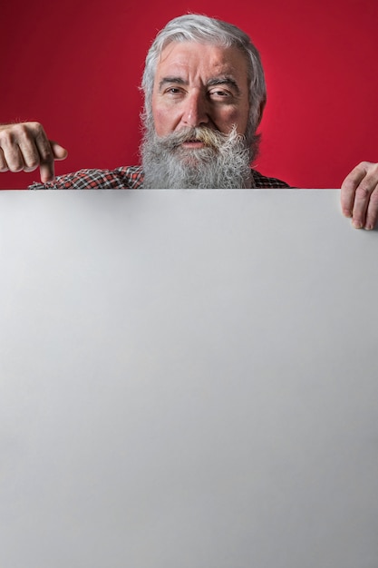 Free photo senior man pointing her finger downward on blank white placard against red background