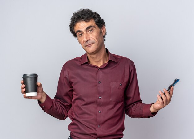 Senior man in purple shirt holdin paper cup and smartphone looking at camera with smile on face standing over white