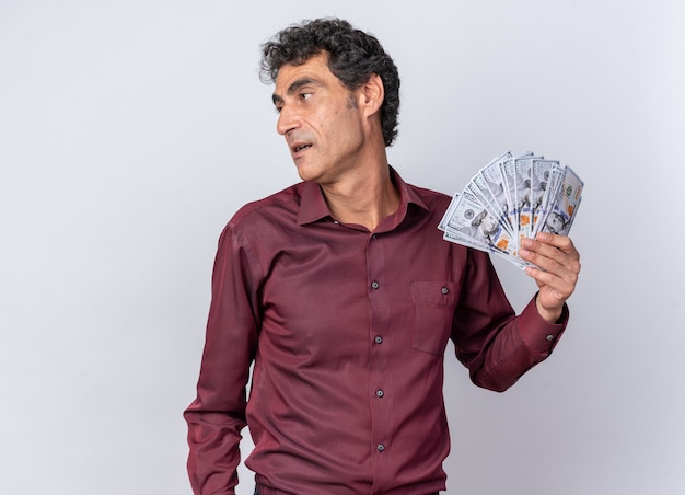 Senior man in purple shirt holding cash looking aside happy and confident standing over white