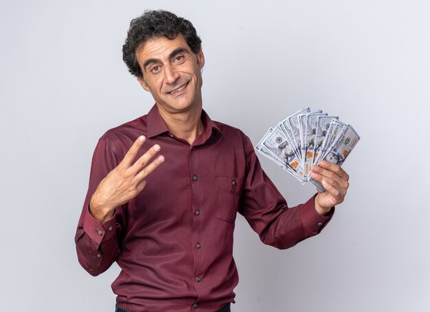 Senior man in purple shirt holding cash looking at camera happy and confident showing number three