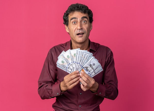 Senior man in purple shirt holding cash looking at camera happy and excited standing over pink background