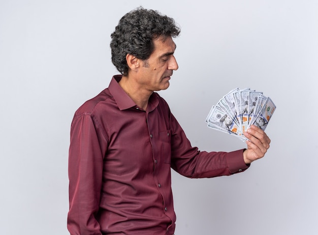 Старший мужчина в фиолетовой рубашке держит деньги, глядя на деньги с серьезным лицом