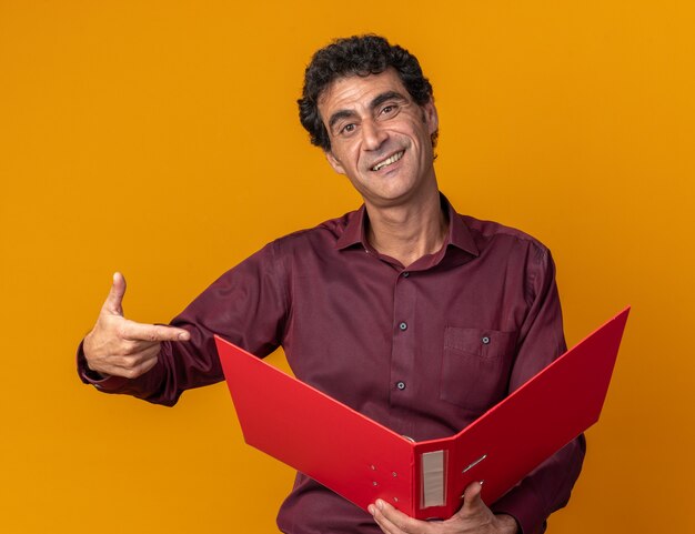 Senior man in purple shirt holding folder pointing with index finger at it smiling confient looking at camera standing over orange