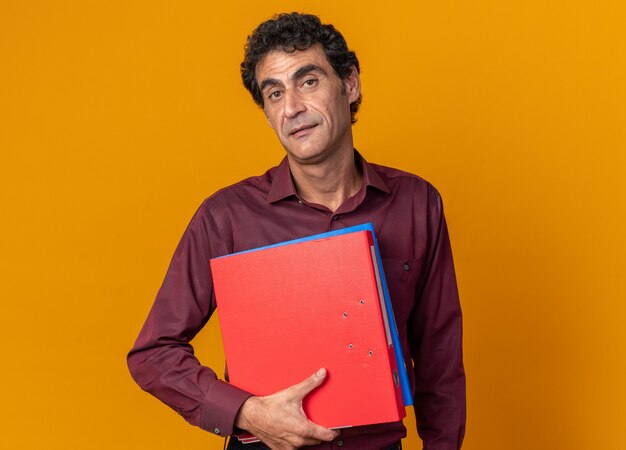 Senior man in purple shirt holding folders looking at camera with serious confident expression standing over orange background