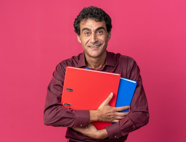 Senior man in purple shirt holding folders looking at camera with smile on happy face standing over pink background