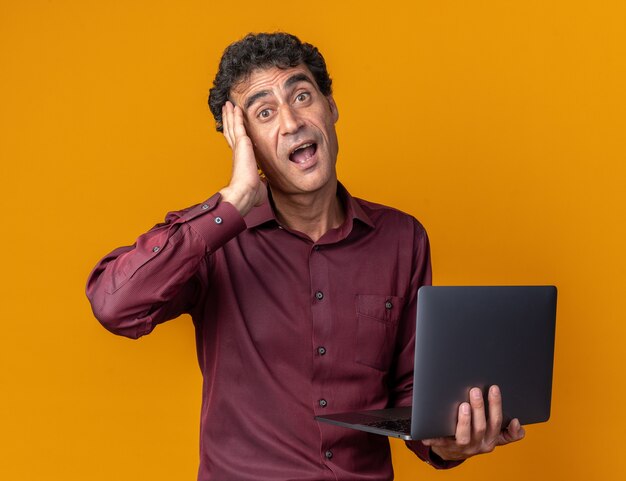 Senior man in purple shirt holding laptop looking at camera confused and surprised with hand on his head standing over orange background