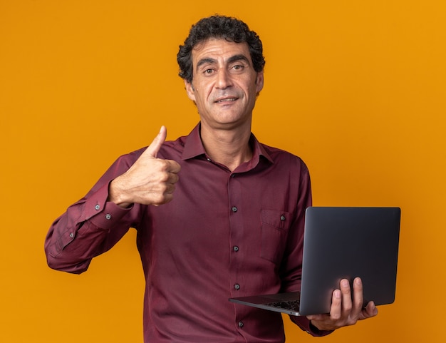 Senior man in purple shirt holding laptop looking at camera smiling confident showing thumbs up standing over orange