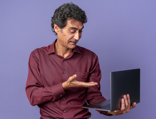 Senior man in purple shirt holding laptop pointing with arm at screen looking confused 