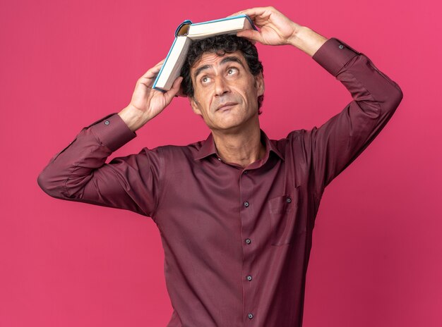 Старший мужчина в фиолетовой рубашке, держащий открытую книгу над головой, выглядит усталым и скучающим, стоя над розовым