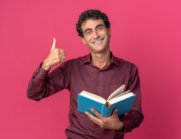 Senior man in purple shirt holding open book looking at camera smiling cheerfully showing thumbs up standing over pink background