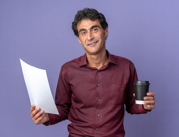 Senior man in purple shirt holding paper cup and blank page looking at camera with smile on face standing over blue background