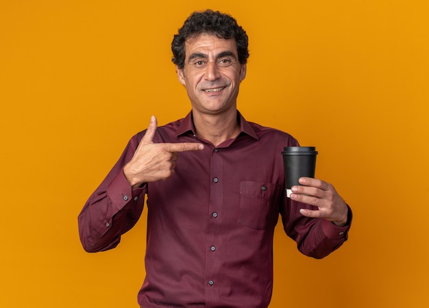 Senior man in purple shirt holding paper cup pointing with index finger at it smiling confident 