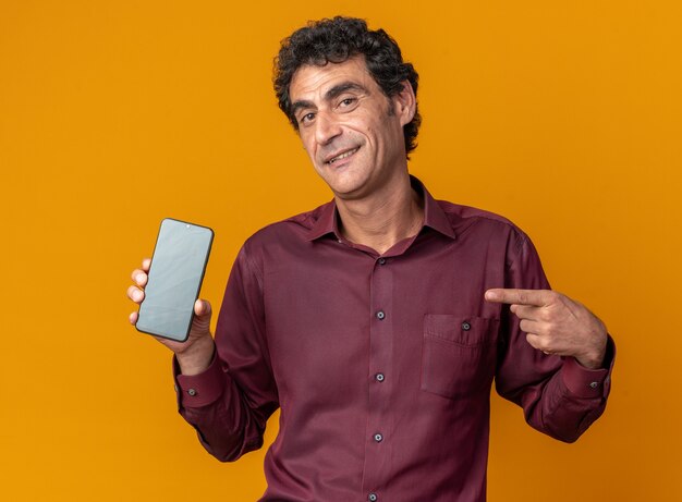 Senior man in purple shirt holding smartphone pointing with index finger at it smiling confident looking at camera standing over orange background