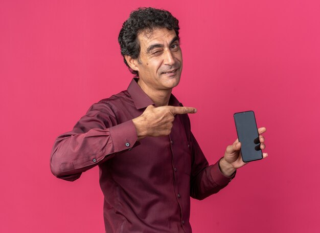 Старший мужчина в фиолетовой рубашке держит смартфон, указывая на него указательным пальцем, улыбается и подмигивает, стоя над розовым