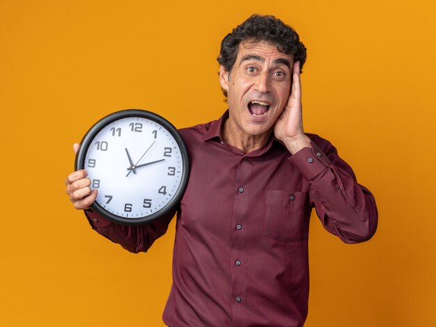 Senior man in purple shirt holding wall clock looking at camera amazed and surprised standing over orange background