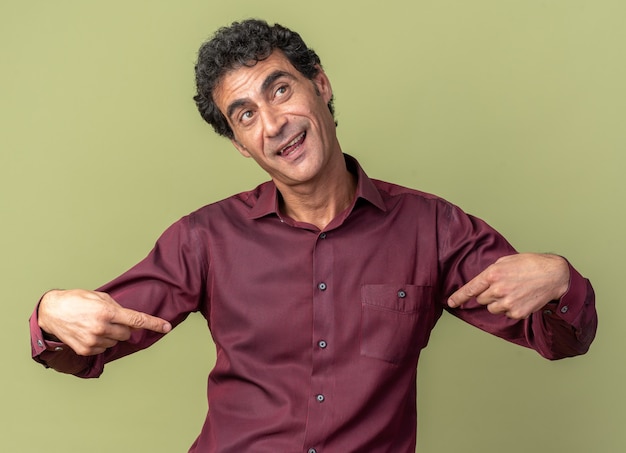 Senior man in purple shirt looking aside happy and cheerful pointing at himself standing over green