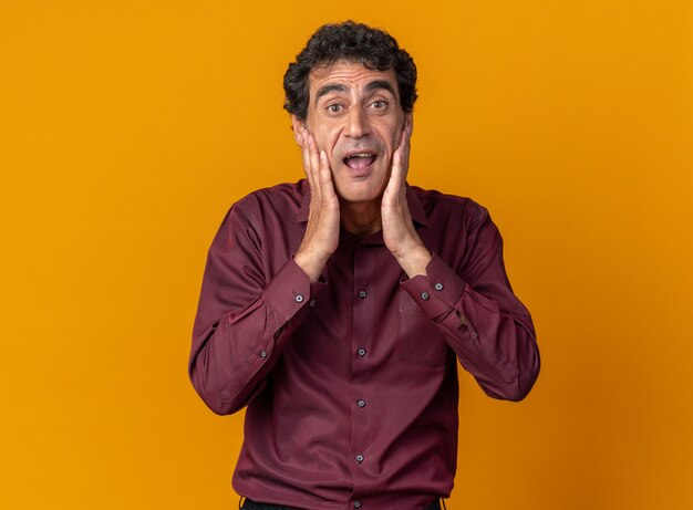 Senior man in purple shirt looking at camera amazed and happy standing over orange background