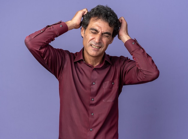 Senior man in purple shirt looking at camera confused and very anxious with hands on his head standing over blue background