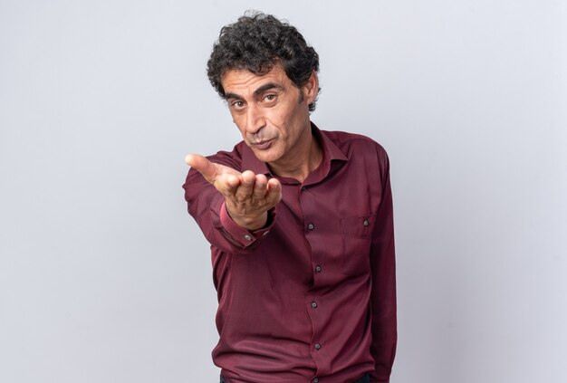 Senior man in purple shirt looking at camera happy and cheerful with hand in front of him blowing a kiss standing over white background