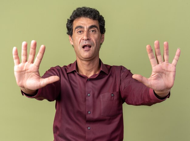 Senior man in purple shirt looking at camera scared making stop gesture with hands holding hands out standing over green