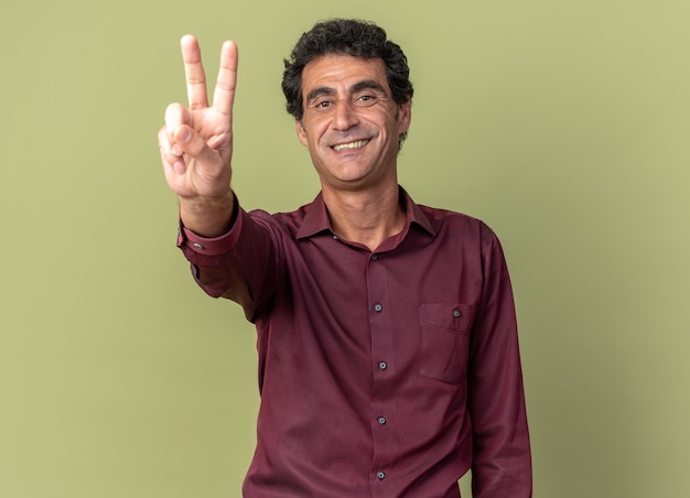 Foto gratuita uomo anziano in camicia viola che guarda l'obbiettivo sorridendo allegramente mostrando il v-sign