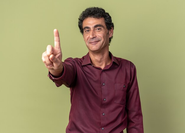 Senior man in purple shirt looking at camera smiling confident showing indsex finger standing over green