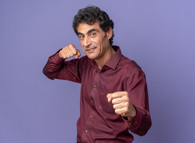 Старший мужчина в фиолетовой рубашке смотрит в камеру со сжатыми кулаками, позирует, как боксер, счастливый и веселый, стоящий над синим