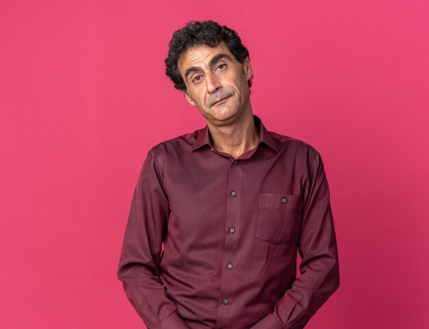 Senior man in purple shirt looking at camera with sad expression on face standing over pink