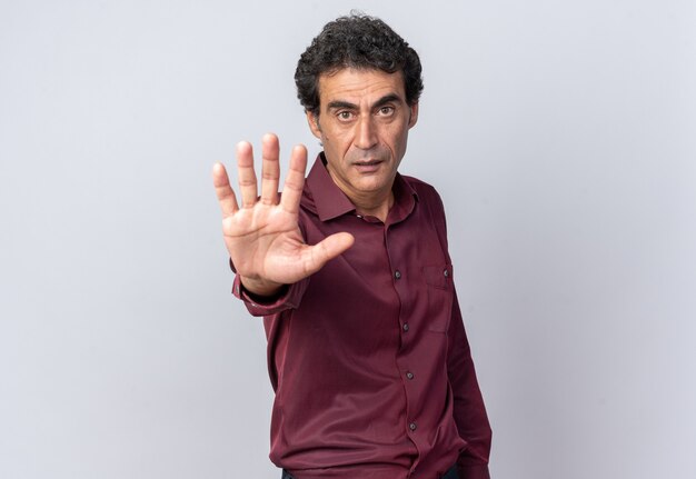 Senior man in purple shirt looking at camera with serious face making stop gesture with hand standing over white background