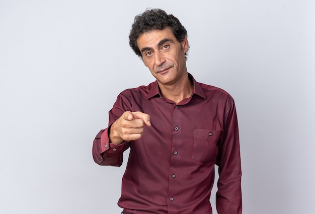 Free photo senior man in purple shirt looking at camera with serious face pointing with index finger at camera standing over white background