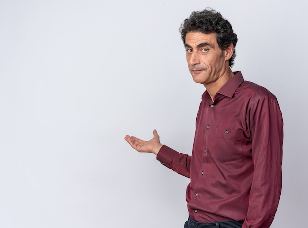 Senior man in purple shirt looking at camera with serious face presenting copy space with arm of his hand standing over white background