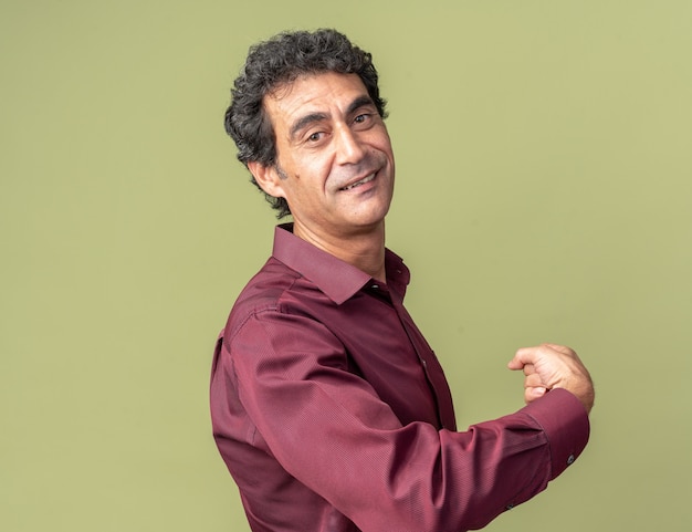 Senior man in purple shirt looking confident smiling pointing back standing over green