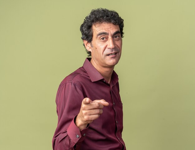 Senior man in purple shirt looking confident smiling pointing with index finger at camera standing over green