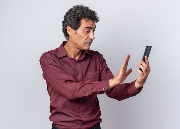 Free photo senior man in purple shirt looking confused holding smartphone making defense gesture