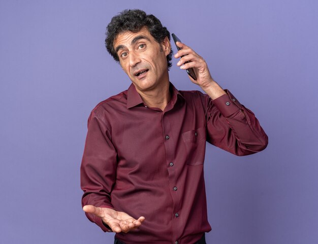 Senior man in purple shirt looking confused while talking on mobile phone standing over blue background