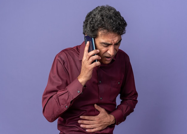 Senior man in purple shirt looking unwell touching his belly while talking on mobile phone 