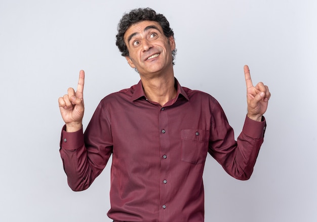 Senior man in purple shirt looking up happy and surprised pointing with index fingers up standing over white background