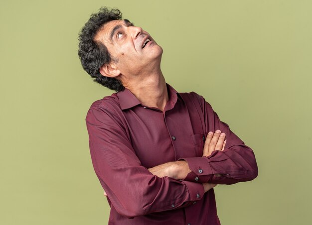 Senior man in purple shirt looking up intrigued with arms crossed standing over green background