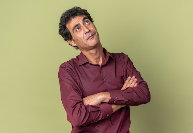 Senior man in purple shirt looking up puzzled with arms crossed standing over green background